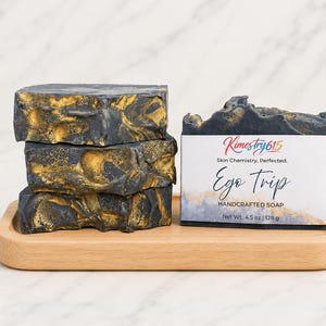 May include: A stack of three rectangular bars of black and gold marbled soap next to a single bar wrapped in a white label. The label reads Kimestry615, Skin Chemistry, Perfected, Ego Trip, HANDCRAFTED SOAP, and Net Wt. 4.5 oz | 128 g.