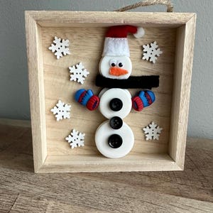 May include: A wooden framed snowman ornament with a white felt hat, black felt scarf, and red felt mittens. The snowman is made of white buttons and has black button eyes and a carrot nose. The background is a wooden surface with white snowflake buttons.
