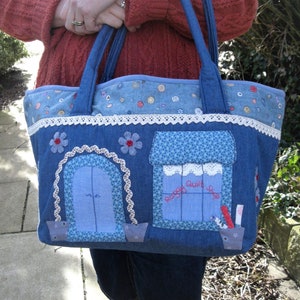 May include: A blue denim tote bag with a patchwork design featuring two buildings. One building has a door and the other has a window with the text "Raggy Quilt Shop".