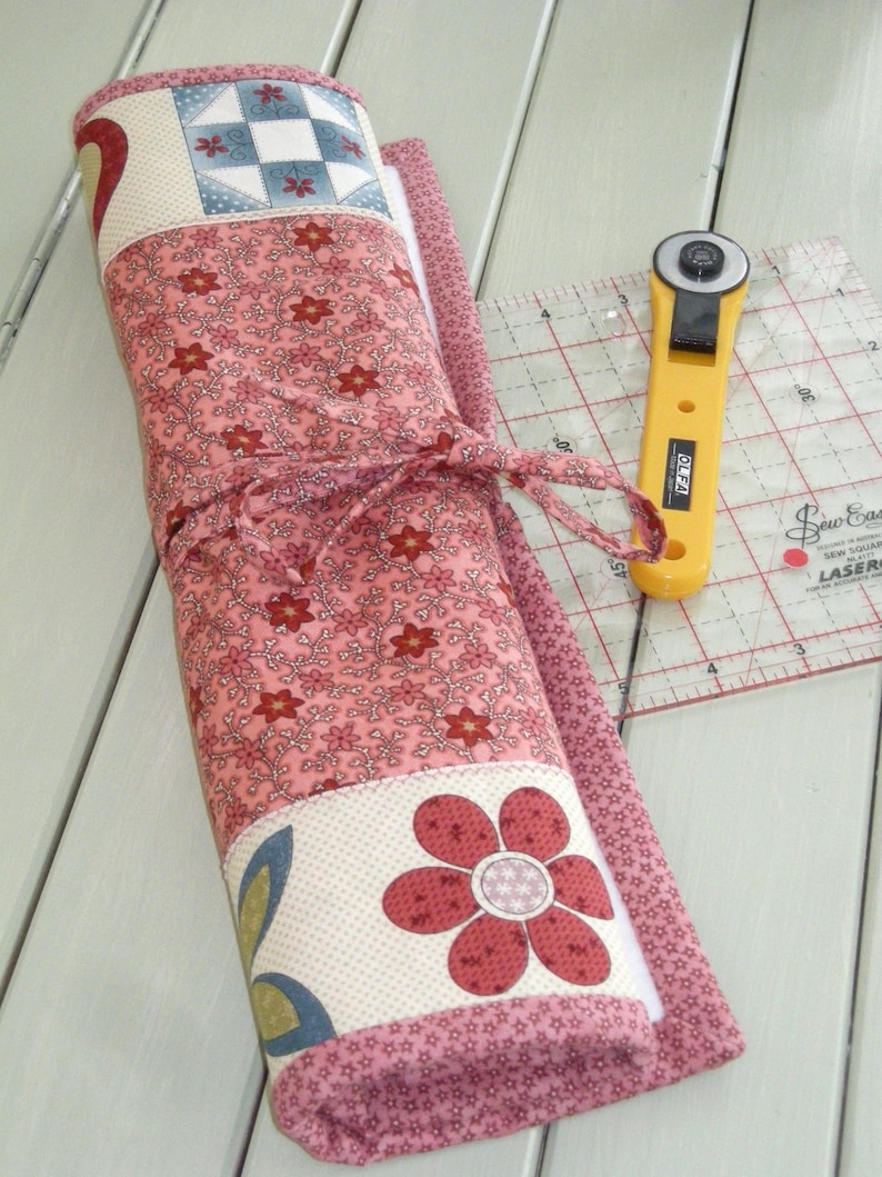 May include: A pink and white fabric roll-up with a floral pattern and a large flower design. The roll-up is tied with a pink ribbon and has a white backing. A yellow rotary cutter is visible in the background.
