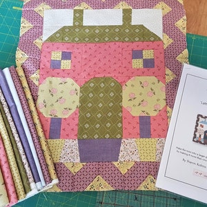 May include: A quilt kit featuring a patchwork design of a house with two trees in front. The kit includes fabric pieces in shades of pink, green, yellow, and purple. The kit is labelled "Lattice Lane" and is described as "Finished".