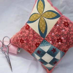 May include: A pink and blue patchwork pincushion with a scalloped edge. The pincushion is filled with pink beads and has a variety of floral patterns.
