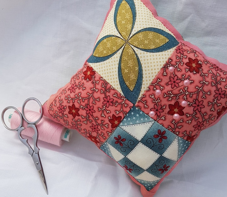 May include: A pink and blue patchwork pincushion with a scalloped edge. The pincushion is filled with pink beads and has a variety of floral patterns.