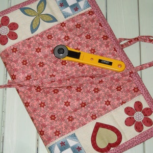 May include: A pink fabric roll-up with a floral pattern and patchwork squares. It has a yellow rotary cutter with the word "OLFA" on it.