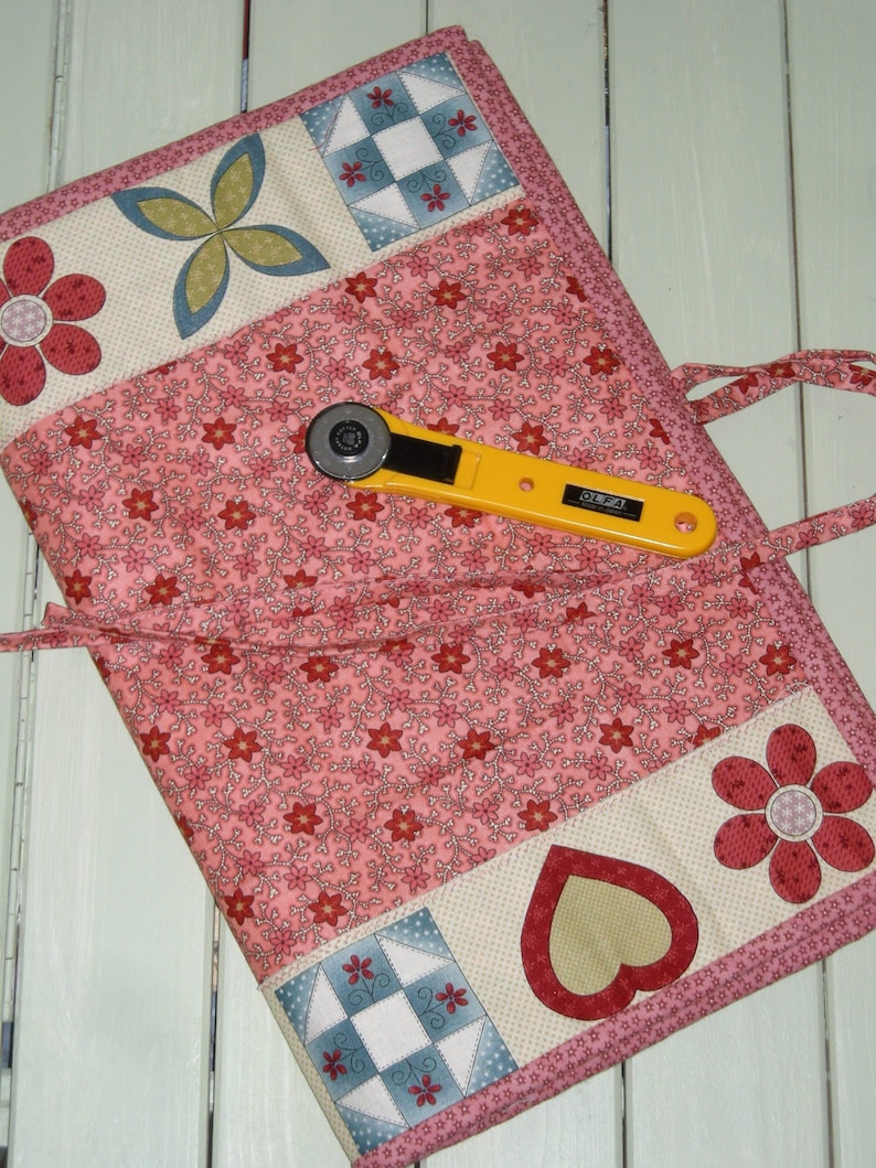 May include: A pink fabric roll-up with a floral pattern and patchwork squares. It has a yellow rotary cutter with the word "OLFA" on it.
