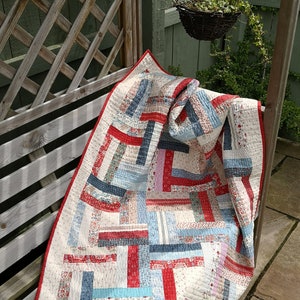 May include: A patchwork quilt with red, blue and white fabric strips in a geometric pattern. The quilt is folded and draped over a wooden bench.