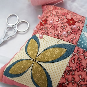 May include: A pink fabric pincushion with a patchwork design featuring floral and geometric patterns. The pincushion is filled with pins with pink heads.