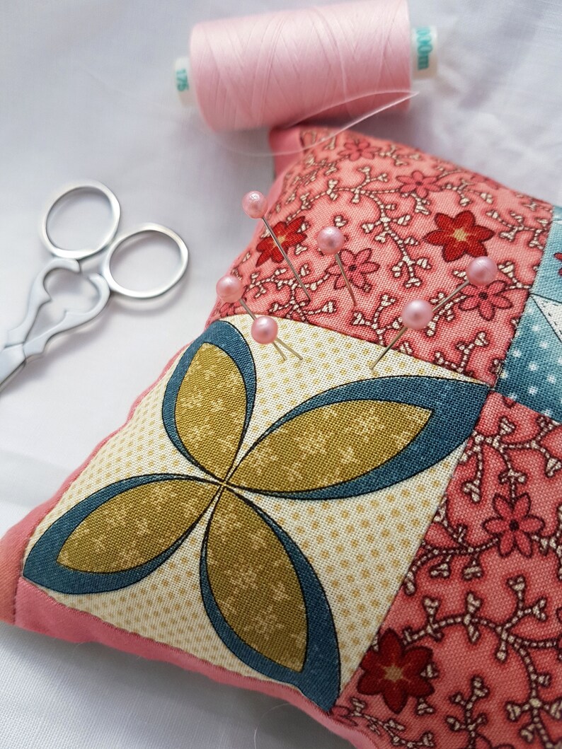 May include: A pink fabric pincushion with a patchwork design featuring floral and geometric patterns. The pincushion is filled with pins with pink heads.
