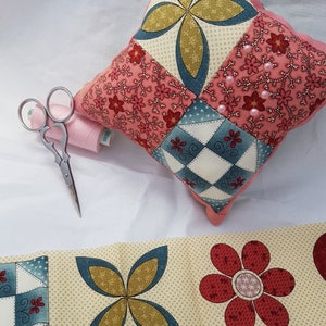 May include: A pink and blue patchwork pincushion with a floral design and pearl accents. The pincushion is sitting on a white surface with a spool of thread and a pair of scissors.