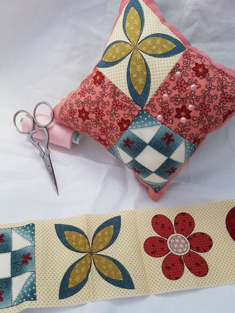 May include: A pink and blue patchwork pincushion with a floral design and pearl accents. The pincushion is sitting on a white surface with a spool of thread and a pair of scissors.
