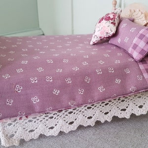 May include: A miniature dollhouse bed with a pink floral patterned bedspread, a pink and white checked pillow, and a floral patterned pillow. The bed has a white crocheted bed skirt.