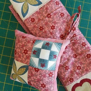 May include: Two quilted fabric squares, one larger than the other, with a pink floral pattern. The smaller square is a pillow with a patchwork design featuring a blue and white checkered pattern, a green and gold butterfly, and a pink floral pattern. The larger square is a runner with a pink floral pattern and a red heart in the corner.
