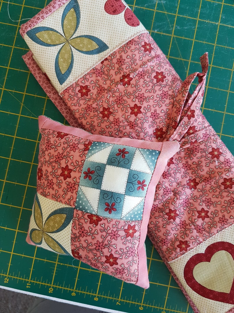 May include: Two quilted fabric squares, one larger than the other, with a pink floral pattern. The smaller square is a pillow with a patchwork design featuring a blue and white checkered pattern, a green and gold butterfly, and a pink floral pattern. The larger square is a runner with a pink floral pattern and a red heart in the corner.