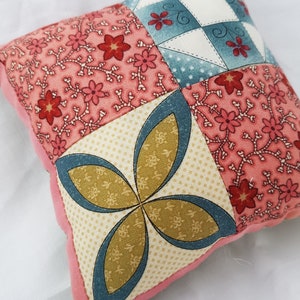 May include: A small pink and cream patchwork pillow with a floral pattern and a four-leaf clover design in the center.