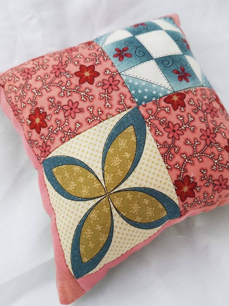May include: A small pink and cream patchwork pillow with a floral pattern and a four-leaf clover design in the center.