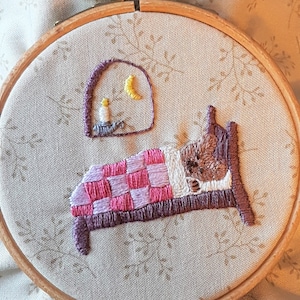 May include: Embroidered hoop art featuring a brown animal sleeping in a pink and purple checked bed with a yellow moon and a lit candle in the background.
