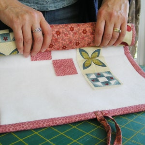 May include: A white fabric roll-up pouch with a pink floral trim and a variety of colorful fabric squares attached. The pouch is rolled up and being held by a person's hands.