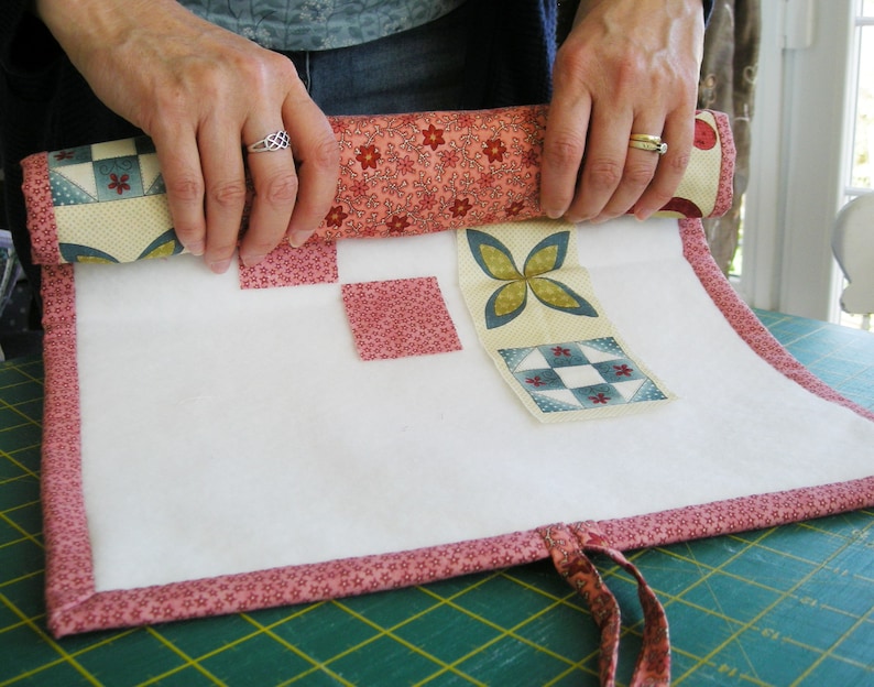 May include: A white fabric roll-up pouch with a pink floral trim and a variety of colorful fabric squares attached. The pouch is rolled up and being held by a person's hands.