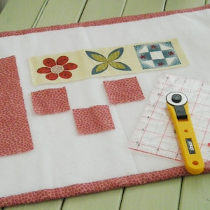 May include: A white fabric surface with pink trim, featuring a row of fabric squares with floral and geometric patterns. There are also several smaller pink fabric squares and a long pink fabric strip. A yellow rotary cutter and a clear ruler with a grid pattern are also visible.