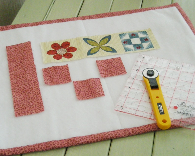 May include: A white fabric surface with pink trim, featuring a row of fabric squares with floral and geometric patterns. There are also several smaller pink fabric squares and a long pink fabric strip. A yellow rotary cutter and a clear ruler with a grid pattern are also visible.