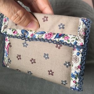 Op de afbeelding: Een kleine, beige stoffen portemonnee met een bloemenpatroon van roze, blauwe en bruine bloemen. De randen zijn afgewerkt met blauw gehaakt kant. De portemonnee wordt in een hand gehouden.