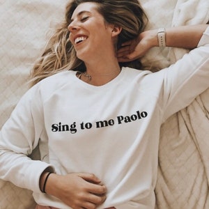 May include: A white long-sleeved sweatshirt with the text "Sing to me Paolo" printed in black on the front.