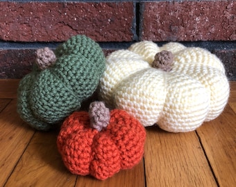 May include: Three crocheted pumpkins in different sizes and colors: green, orange, and white. The pumpkins are sitting on a wooden surface.