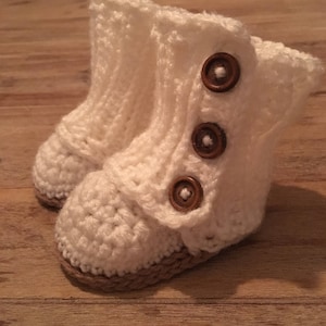 Puede incluir: Un par de botines de bebé de ganchillo, color blanco roto. Los botines tienen un cierre de botón con tres botones marrones. Las suelas son de color marrón claro. Los botines están hechos de hilo suave y texturizado.