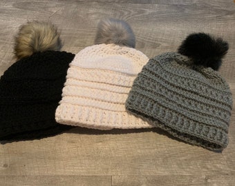May include: Three crocheted hats with faux fur pom-poms. The hats are black, white, and gray.