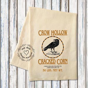 K&ouml;nnte beinhalten: Ein beigefarbenes K&uuml;chentuch mit einer schwarzen Kr&auml;henillustration und dem Text "CROW HOLLOW CRACKED CORN CROW HOLLOW MILLING CO. CROW HOLLOW, PA 50 LBS. NET WT."