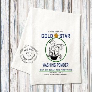 May include: A white tea towel with a vintage-style illustration of a girl doing laundry. The text on the towel reads "5 LBS. NET WT. GOLD STAR WASHING POWDER GET 'EM CLEAN THE FIRST TIME DISTRIBUTED BY: GOLD STAR SOAP COMPANY". There is also a small stamp that reads "etsy shop Antique Primitives".