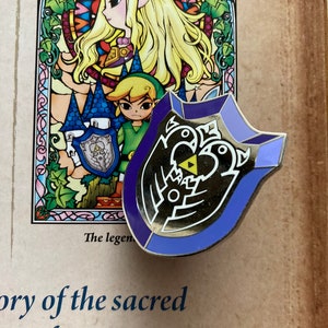 May include: A purple and silver enamel pin featuring the Triforce symbol from the Legend of Zelda video game series. The pin is shaped like a shield and has a detailed design.