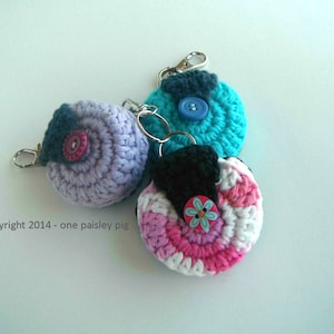May include: Three crocheted keychains with different colors and button embellishments. The keychains are turquoise, purple, and pink with white accents. Each keychain has a silver metal ring.