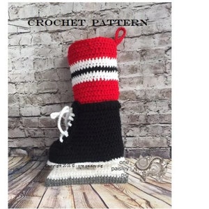 Puede incluir: Patrón de crochet para una media de hockey sobre hielo roja, blanca y negra. La media tiene una bota negra con un cordón blanco, un puño rojo con rayas blancas y una suela gris.