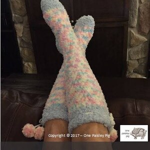 May include: A pair of cozy, fluffy reading socks in a pastel pink, blue, and white color scheme. The socks have a pom-pom detail at the top.