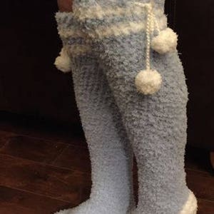 May include: A pair of blue and white fuzzy knee-high socks with white pom-poms. The socks have a white band around the top and a white trim at the bottom. The socks are called "Coziest Reading Socks" and are a crochet pattern by One Paisley Pig.