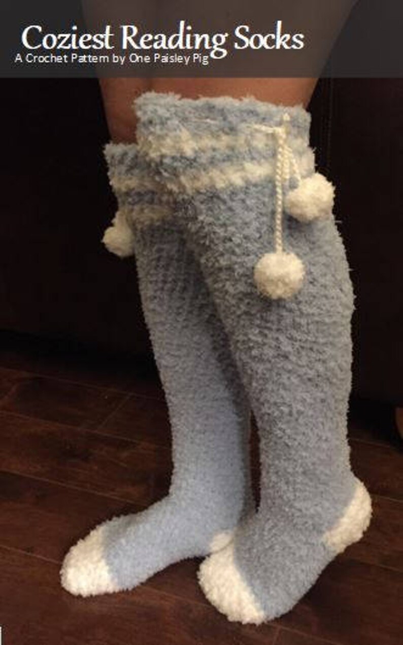May include: A pair of blue and white fuzzy knee-high socks with white pom-poms. The socks have a white band around the top and a white trim at the bottom. The socks are called "Coziest Reading Socks" and are a crochet pattern by One Paisley Pig.