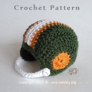 Peut inclure: Chapeau en forme de casque de football américain crocheté en vert, orange et blanc. Le chapeau a une sangle blanche et un bouton sur le côté. Le texte "Copyright 2015 - One Paisley Pig" est en bas de l'image.