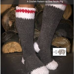 May include: A pair of gray crochet reading socks with white and red stripes at the top. The socks are sitting on a pile of logs. The text "Coziest Reading Socks" and "A Crochet Pattern by One Paisley Pig" is at the top of the image. The text "Copyright &copy; 2017 - One Paisley Pig" is at the bottom of the image.