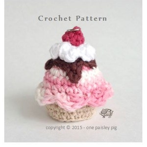 Könnte beinhalten: Ein gehäkelter Eiswaffelkegel aus rosa, weißem und braunem Garn. Der Eiswaffelkegel hat eine rote Kirsche obenauf und einen braunen Schokoladensoßen-Drizzle. Das Häkelmuster stammt von One Paisley Pig.