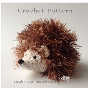 Könnte beinhalten: Häkelanleitung für ein braunes und weißes Igel-Amigurumi-Spielzeug. Der Igel hat einen flauschigen braunen Körper und ein weißes Gesicht mit schwarzen Augen und einer Nase.