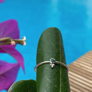 Könnte beinhalten: Ein silberner Ring mit einem kleinen, runden, grünen Edelstein und drei kleineren silbernen Perlen. Der Ring wird auf einem grünen Blatt mit lila Blumen und einem blauen Hintergrund gezeigt.