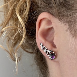 Könnte beinhalten: Ein silberner Ohrring mit einem violetten Edelstein. Der Ohrring ist mit komplizierten Wirbeln und Kurven verziert.