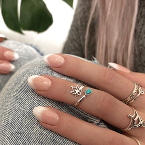 Könnte beinhalten: Nahaufnahme von Händen mit weißen, mandelförmigen Nägeln. Der Ringfinger trägt einen silbernen Ring mit einem türkisfarbenen Stein und einem Lotusblumen-Design. Der Mittelfinger trägt drei silberne Ringe mit filigranen Designs.