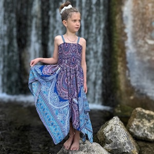 Może przedstawiać: Młoda osoba w sukience bez rękawów we wzory z niebieskimi, fioletowymi i białymi wzorami paisley. Sukienka ma płynny, asymetryczny dół. Osoba stoi na skale w pobliżu wodospadu, z wodospadem w tle.