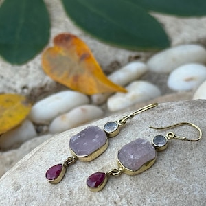 Può includere: Un paio di orecchini color oro con una pietra ovale viola chiaro, una piccola pietra rotonda blu chiaro e una piccola pietra rotonda rossa. Gli orecchini sono esposti su una roccia grigia con foglie verdi e gialle sullo sfondo.