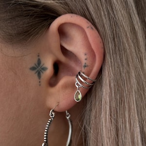 Op de afbeelding: Een zilveren oorring met een gedetailleerd ontwerp en een zilveren oormanchet met een groene edelsteen hanger.