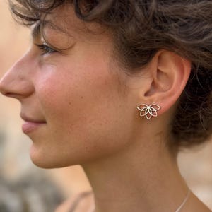Pode incluir: Um brinco de pino metálico com um design floral simétrico e vazado, apresentando seis pétalas arredondadas dispostas em um grupo. O brinco é feito de metal polido de cor clara e é usado no lóbulo da orelha, exibindo uma silhueta delicada.