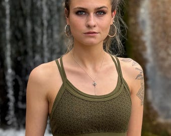 Pode incluir: Uma mulher vestindo um sutiã esportivo verde com um padrão geométrico. O sutiã tem um estilo de costas de nadador e um design de alça dupla. Ela está em pé na frente de uma cachoeira.