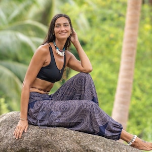 May include: A woman wearing a black sports bra and blue patterned pants sits on a large rock in a tropical setting. She is smiling and has long dark hair.